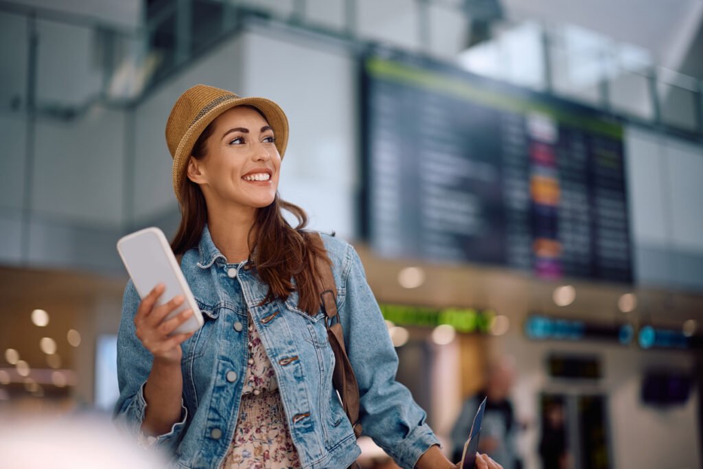 Happy traveler using cell phone while waiting for her flight at the airport.